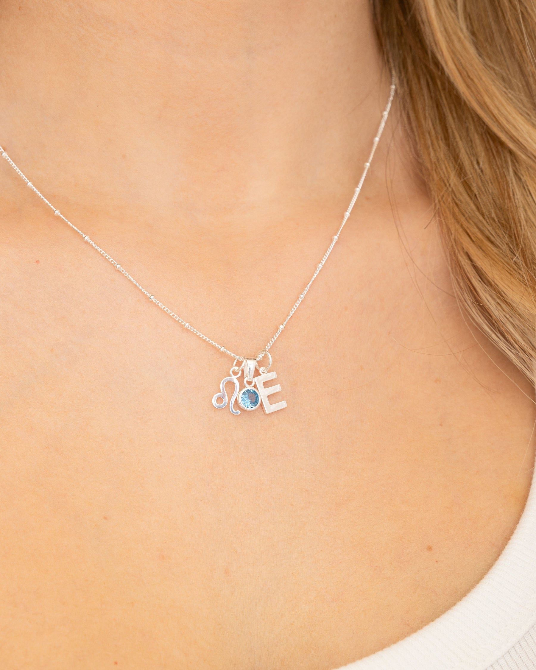 Sterling silver necklace featuring a letter E pendant, a Leo zodiac charm, and a small blue gemstone on a delicate chain.